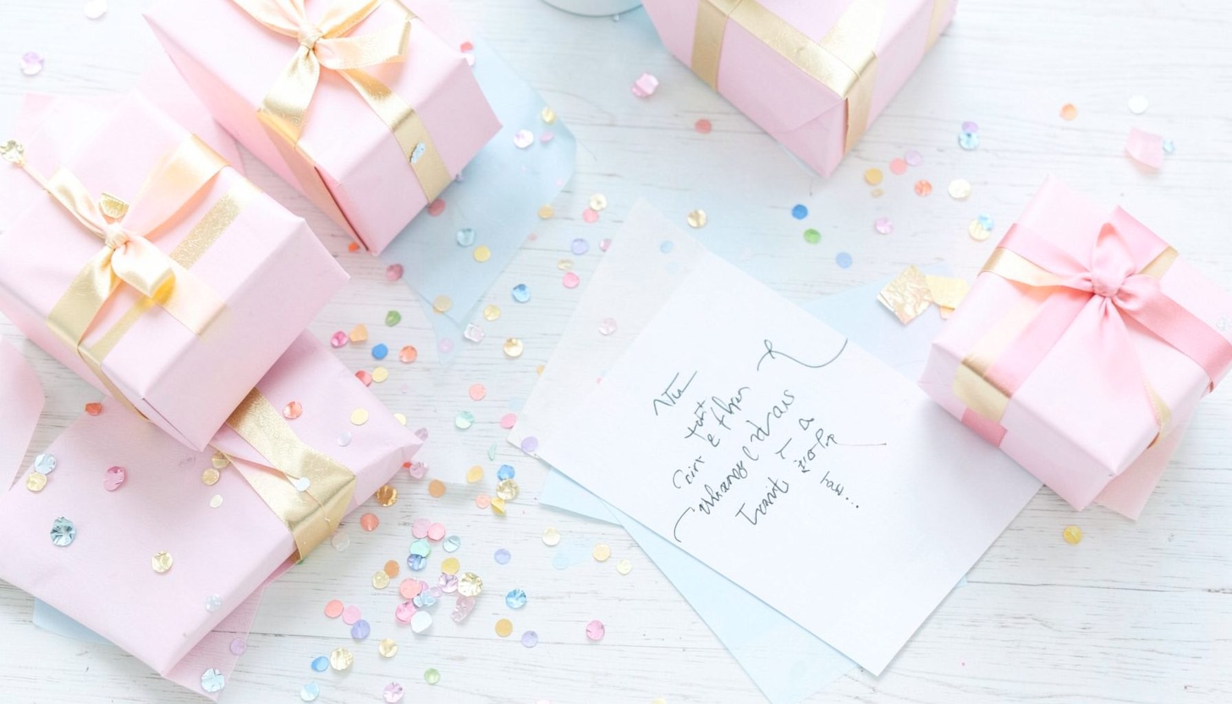 Bright, airy flat lay of wrapped gift boxes in pastel pink and gold tones, scattered confetti, and a handwritten celebration card