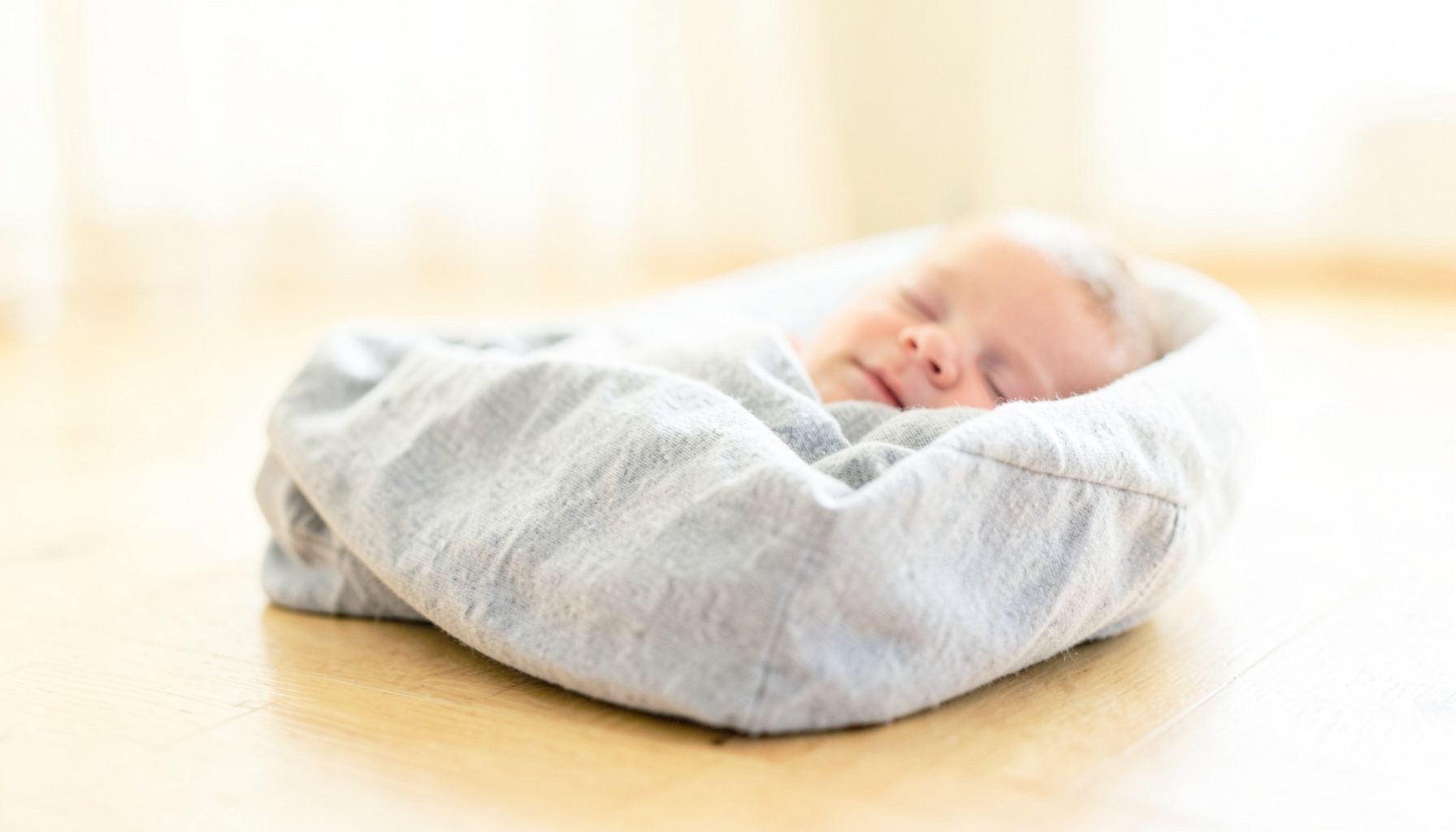 A close-up of a baby sleeper with soft gray fabric and a gentle incline, resting on a light wood floor in a bright, airy room